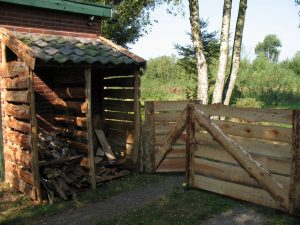 Een toegangshek met een kleine en grote poort. De palen van het hek gaan over in de constructie van het houthok.  